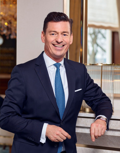 image 10 Ein Mann in einem dunklen Anzug und mit blauer Krawatte steht in einem Haus, lächelt selbstbewusst und stützt sich mit einer Hand auf eine Tischplatte. Im Hintergrund sind ein Fenster und eine dekorative Beleuchtung zu sehen.