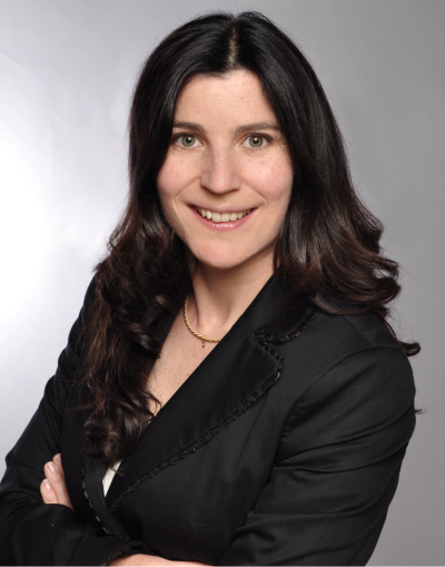 image 8 Eine Frau mit langen, dunklen, gewellten Haaren, die einen schwarzen Blazer und eine goldene Halskette trägt, lächelt in die Kamera vor einem schlichten grauen Hintergrund. Ihre Arme sind verschränkt.