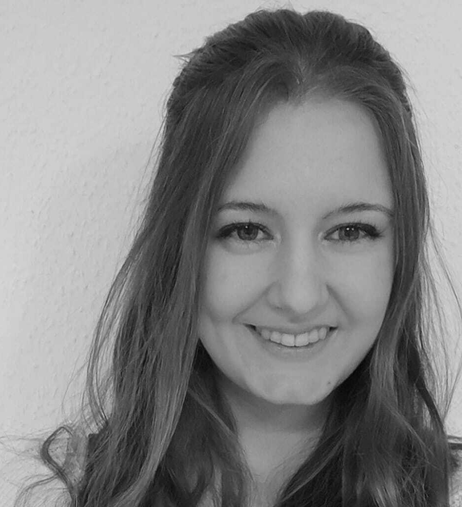 Julia Bittan Schwarz-Weiß-Foto einer jungen Frau mit langen Haaren, die in die Kamera lächelt, vor einem schlichten hellen Hintergrund.