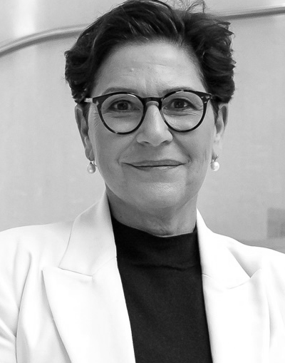 Eine Frau mit kurzen dunklen Haaren, die eine runde Brille, Perlenohrringe, ein dunkles Oberteil und einen hellen Blazer trägt, lächelt leicht, während sie in einem Innenraum steht. Das Foto ist in schwarz-weiß gehalten.