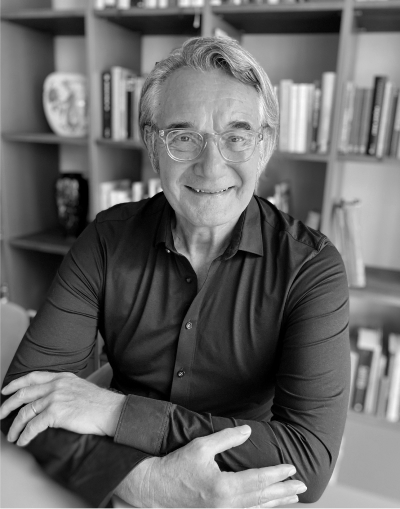 image-14-2 Ein lächelnder älterer Mann mit Brille und grauem Haar sitzt an einem Tisch mit verschränkten Armen vor einem mit Büchern und Dekorationsgegenständen gefüllten Bücherregal. Das Foto ist in schwarz-weiß gehalten.