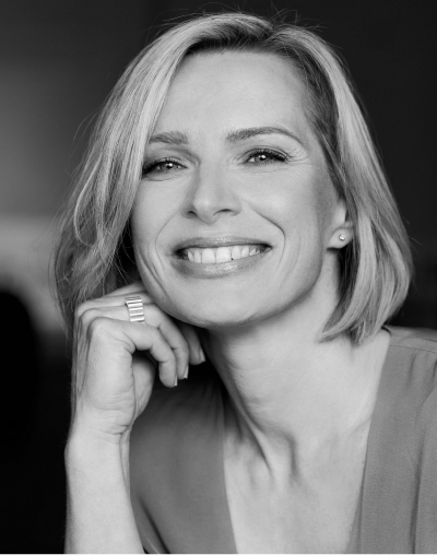 image-6-2 Schwarz-Weiß-Foto einer lächelnden Frau mit kurzen, blonden Haaren, die ein helles Oberteil trägt. Sie stützt ihr Kinn auf ihre Hand und blickt selbstbewusst in die Kamera.