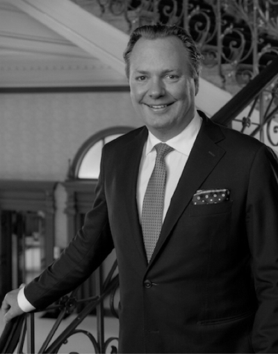 image-9-2 Ein Mann in Anzug und Krawatte steht lächelnd an einer verschnörkelten Treppe, eine Hand auf das Geländer gestützt, in einer eleganten Inneneinrichtung.