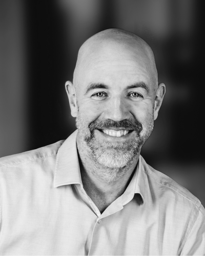 schneider-2 Schwarz-Weiß-Porträt eines Mannes mittleren Alters mit Glatze und Bart, der freundlich in die Kamera lächelt. Er trägt ein helles Hemd mit Kragen, der Hintergrund ist unscharf.