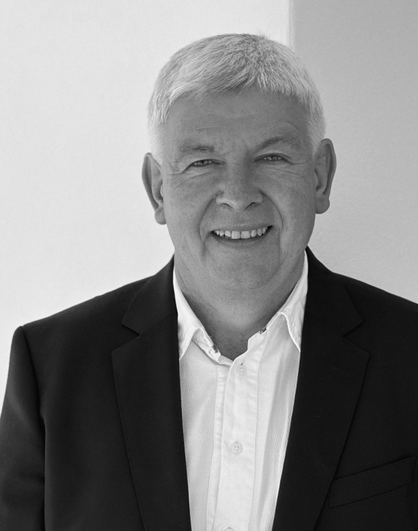 thuy-600×763-2 Ein lächelnder älterer Mann mit kurzen Haaren, der einen dunklen Blazer über einem weißen Button-up-Hemd trägt, steht vor einem schlichten hellen Hintergrund.