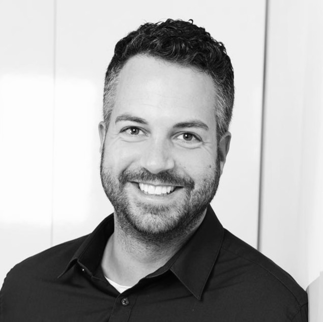 Jan Ulrich Ein Mann mit kurzen lockigen Haaren und einem Bart lächelt in die Kamera. Er trägt ein dunkles Hemd und steht vor einem hellen Hintergrund. Das Foto ist in schwarz-weiß gehalten.