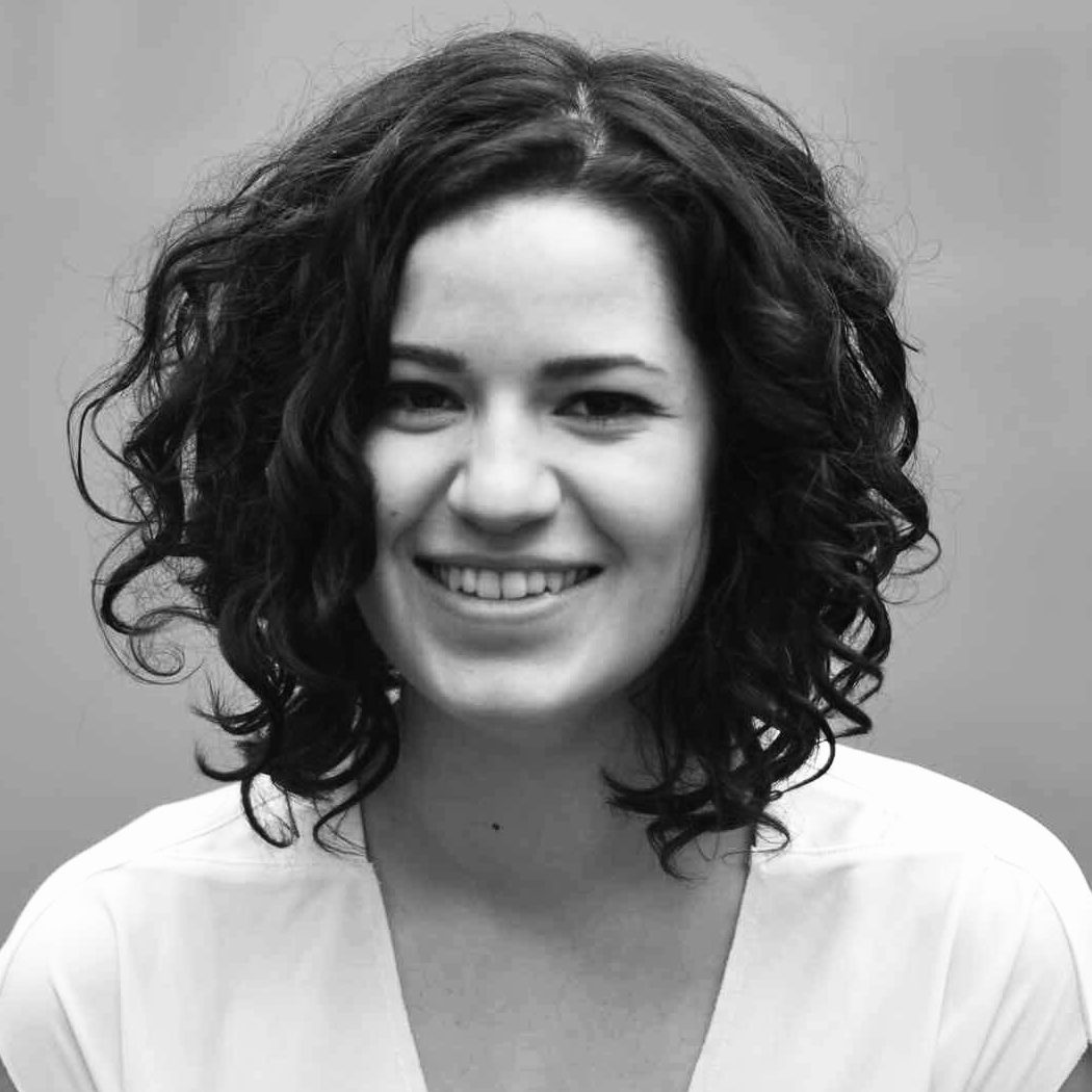 Lara Liese Eine Frau mit lockigem dunklem Haar lächelt in die Kamera. Sie trägt ein helles Oberteil und der Hintergrund ist schlicht und unscharf. Das Bild ist in schwarz-weiß gehalten.